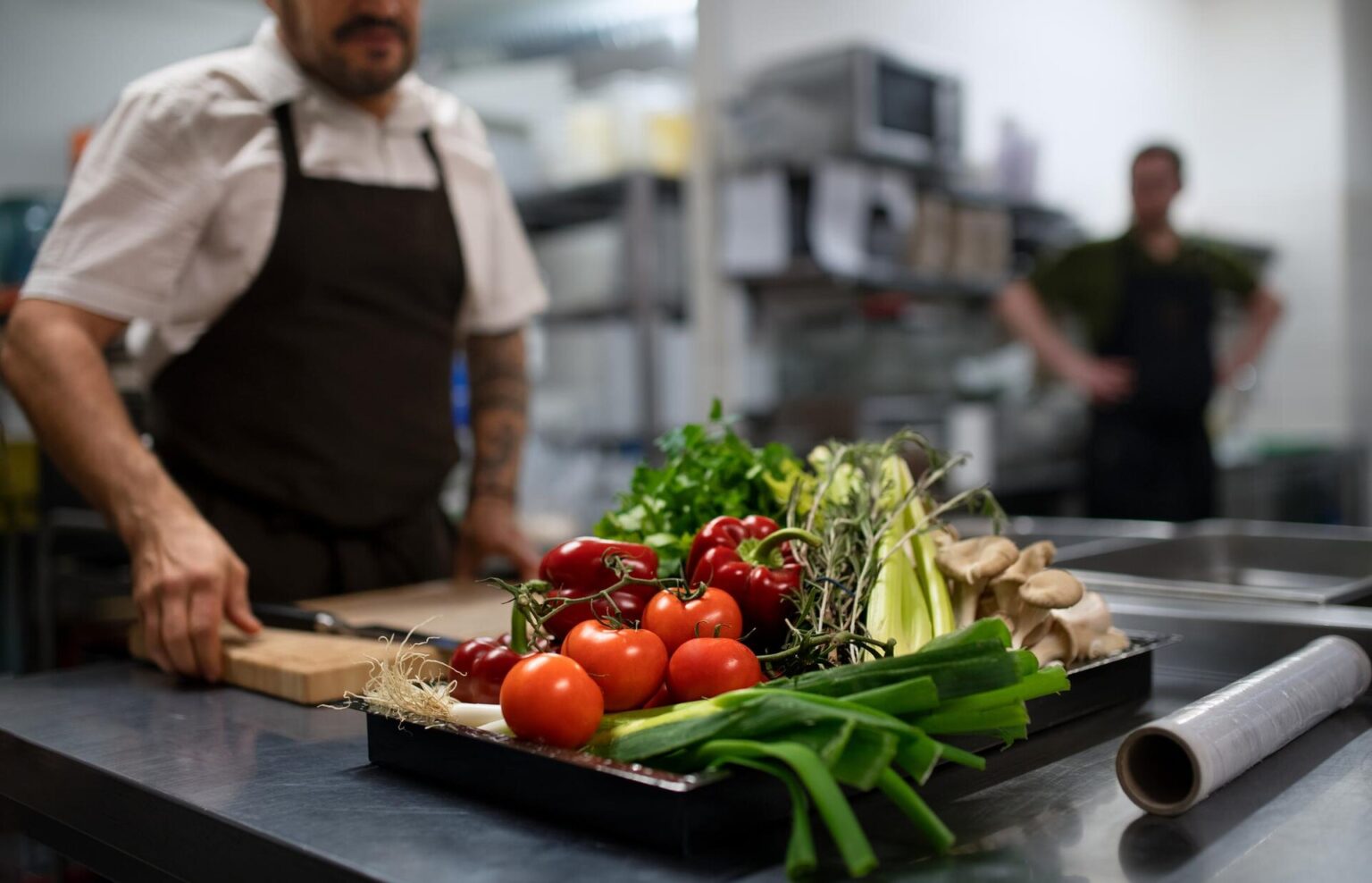 Cozinheiro de avental na bancada de inox com bandeja de tomates, pimentões, cebolinha, ervas e cogumelos